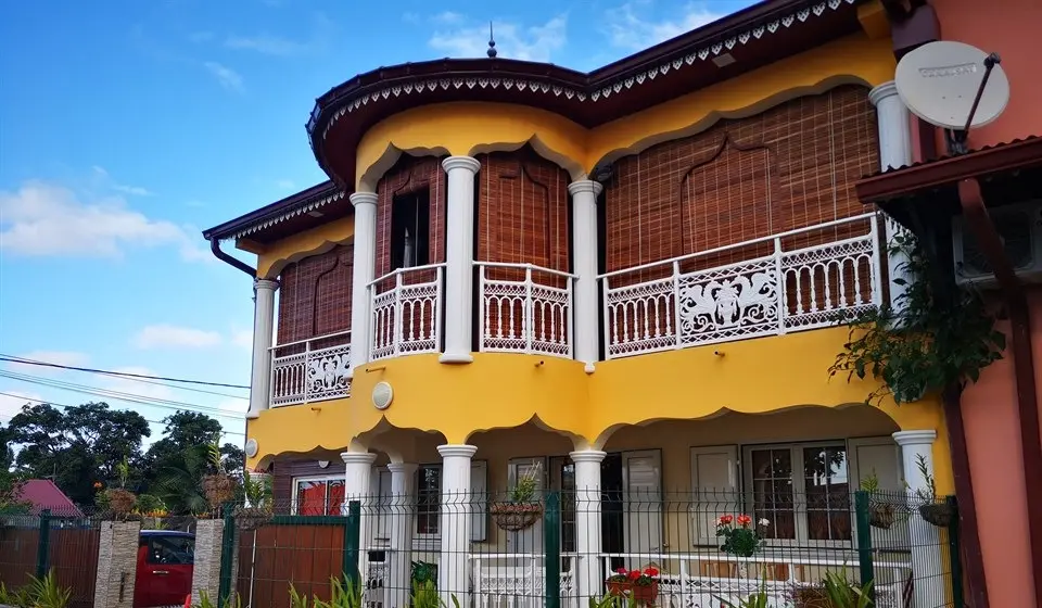 Maison créole jaune à étage dont les varangues sont fermées par des stores mauriciens découpés en pointe